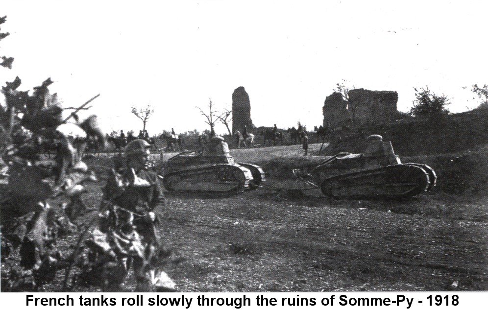Black and white photo of two small French Renault light tanks, one towing the other, rolling along a muddy road with crumbling stoneworks behind them. A single French soldier stands behind foliage in the foreground. Behind the tanks a line of soldiers on horseback moves in the opposite direction from that of the tanks.